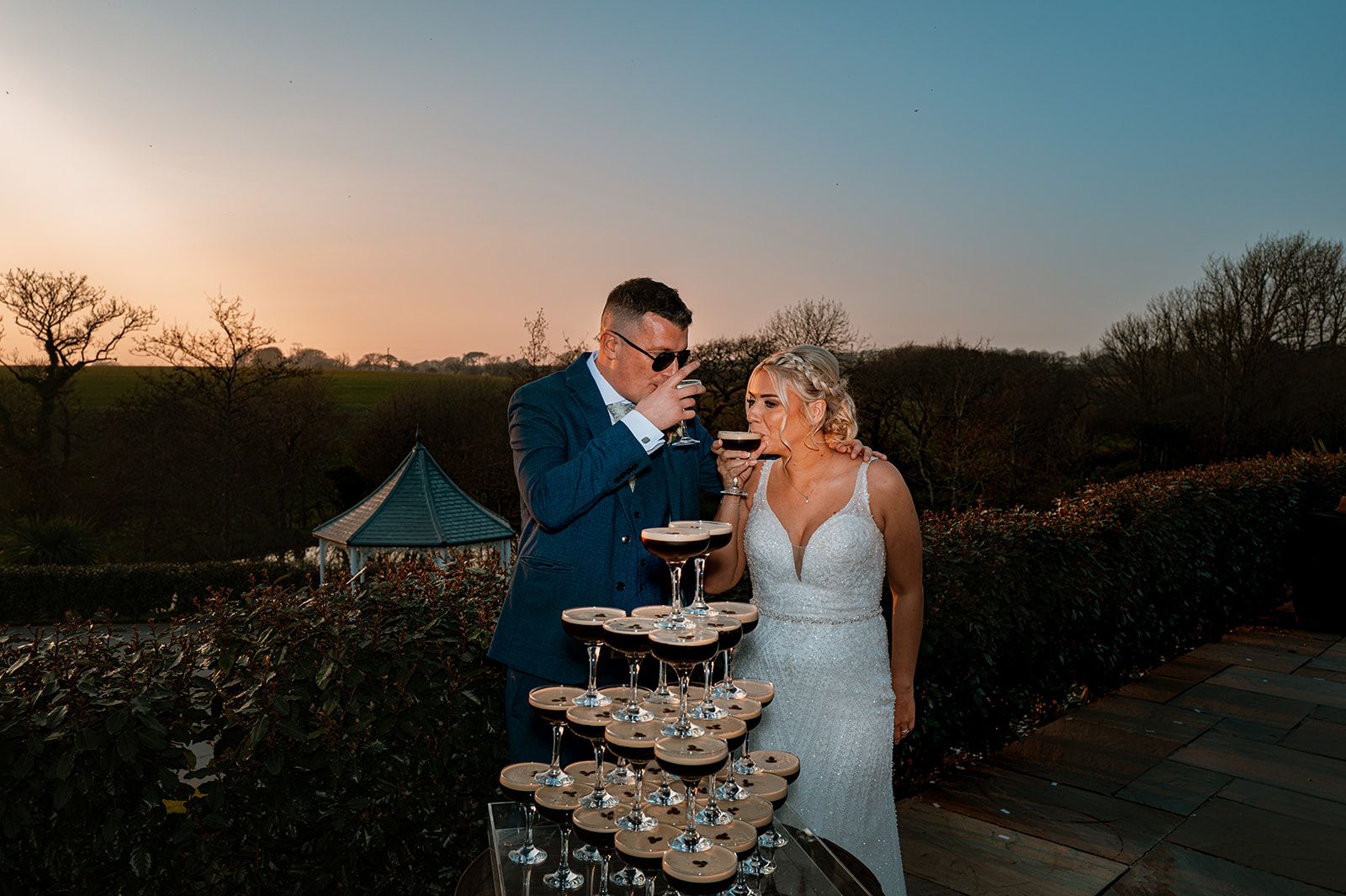 The bride and groom stand outdoors, drinking espresso martinis from a stacked cocktail tower.
