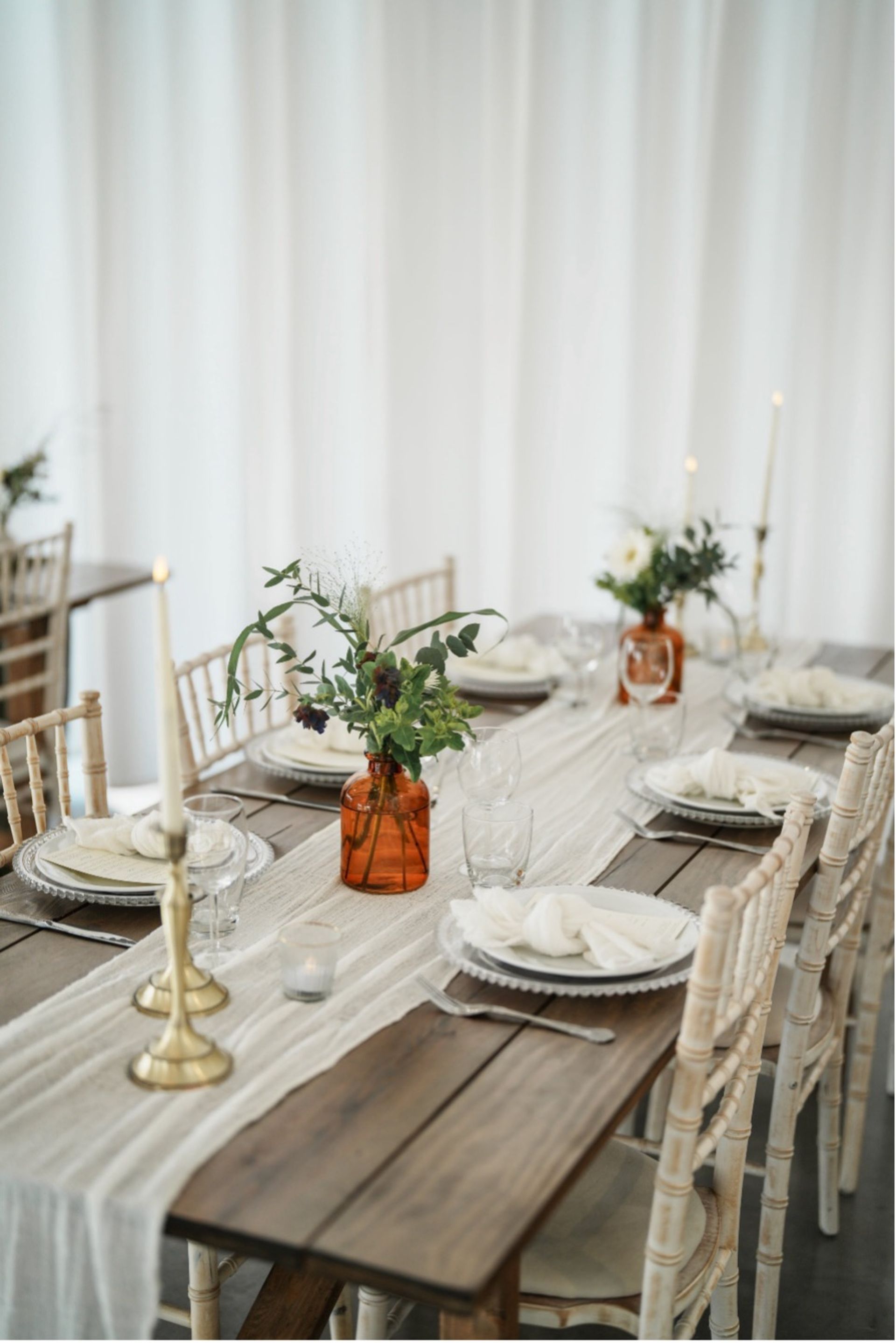 Rustic wooden wedding table decorated with a soft white gauze runner, amber glass bud vases filled with seasonal greenery, brass candlesticks, white place settings and Chiavari chairs.