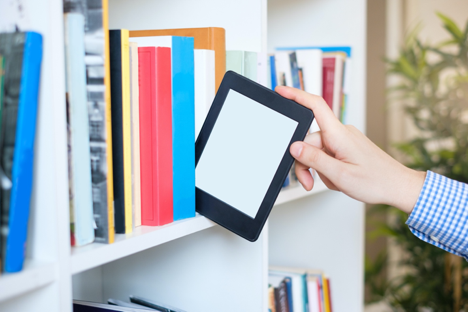 A hand reaches out to pull a e-reader device off a shelf of paper books. 
