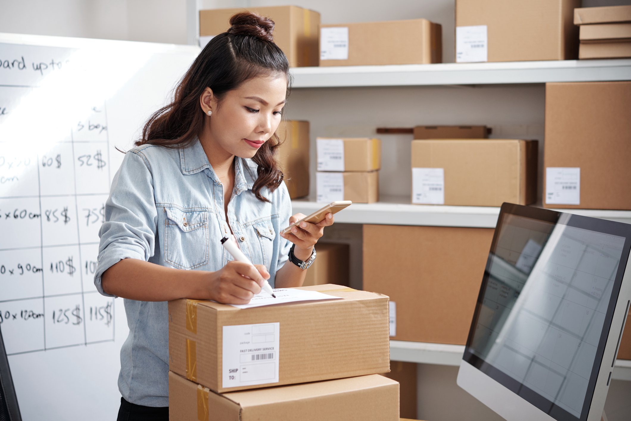 Woman packing product boxes for her business, looking at her cell phone to confirm information,  ecommerce business models