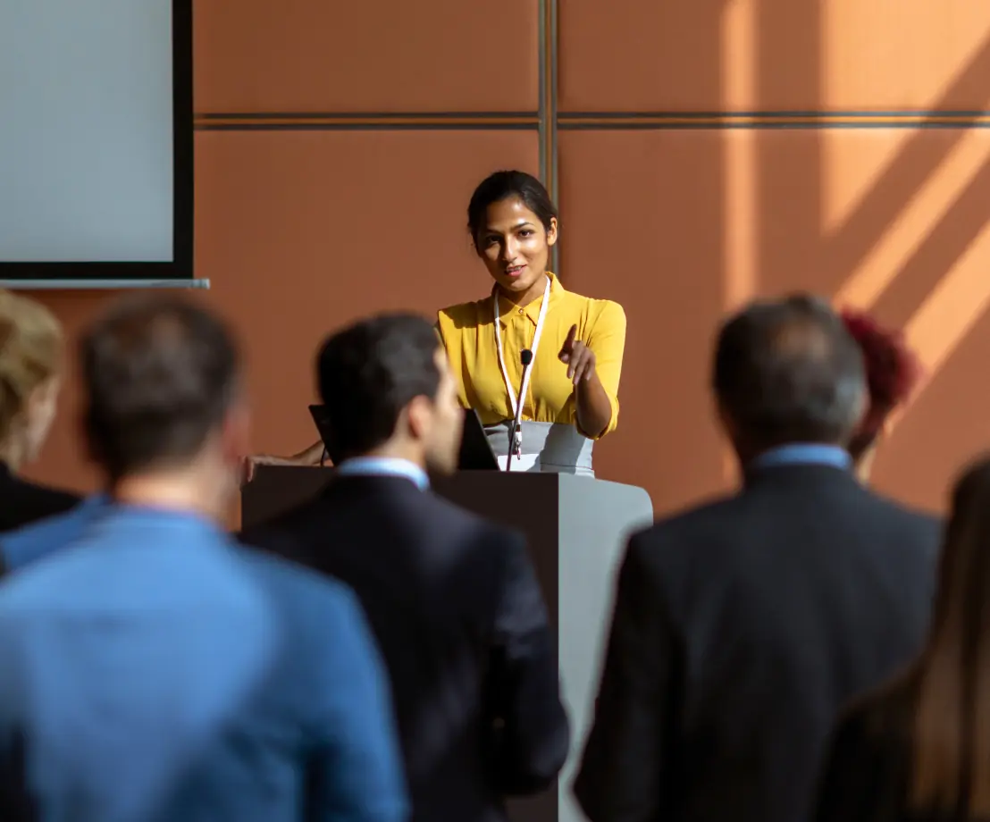 A woman stands behind a podium and addresses a room full of people in business attire.
