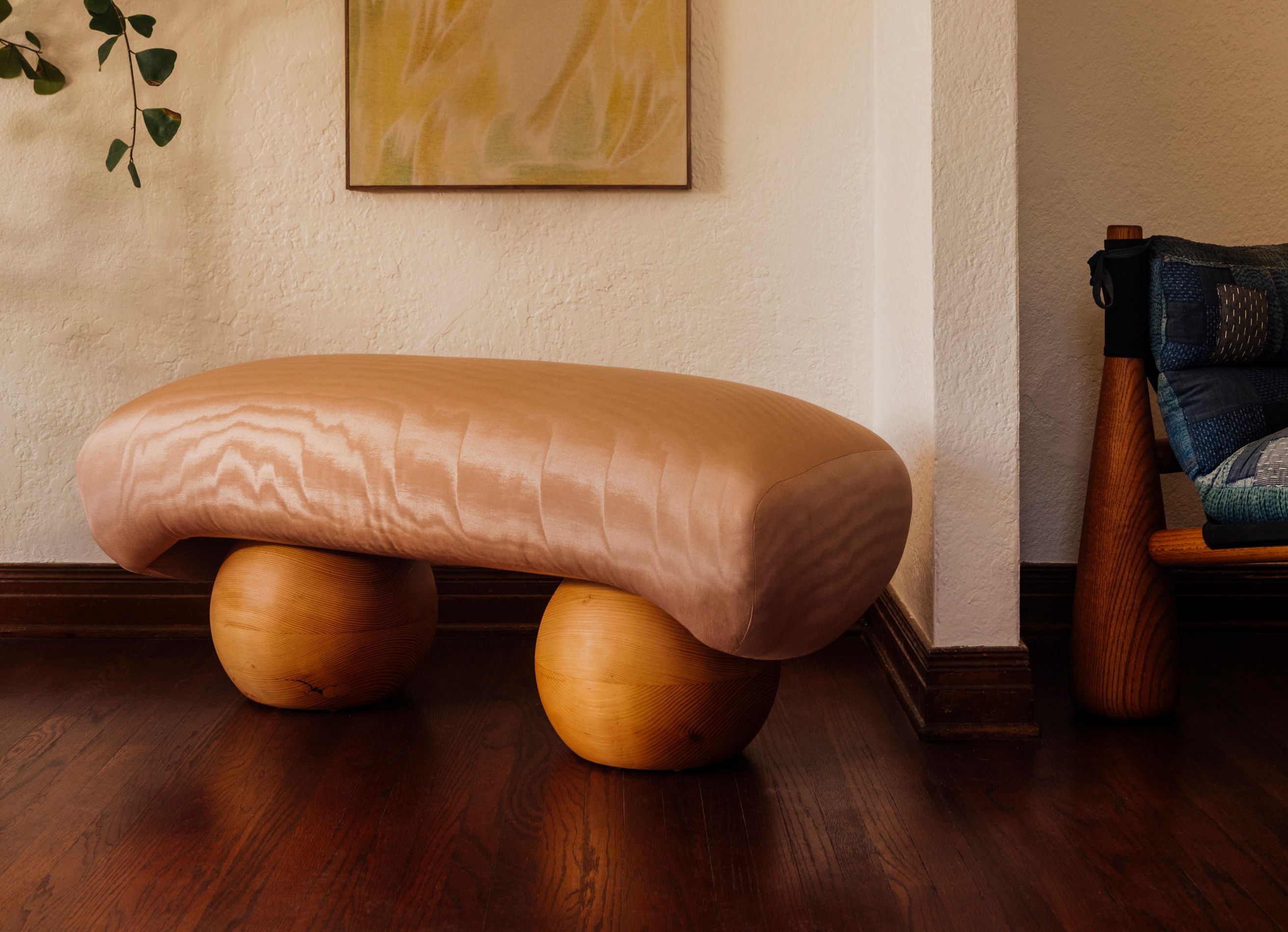 Bench with sphere legs and silky pink fabric sits in a living room with wood floor next to a chair and cream colored walls.