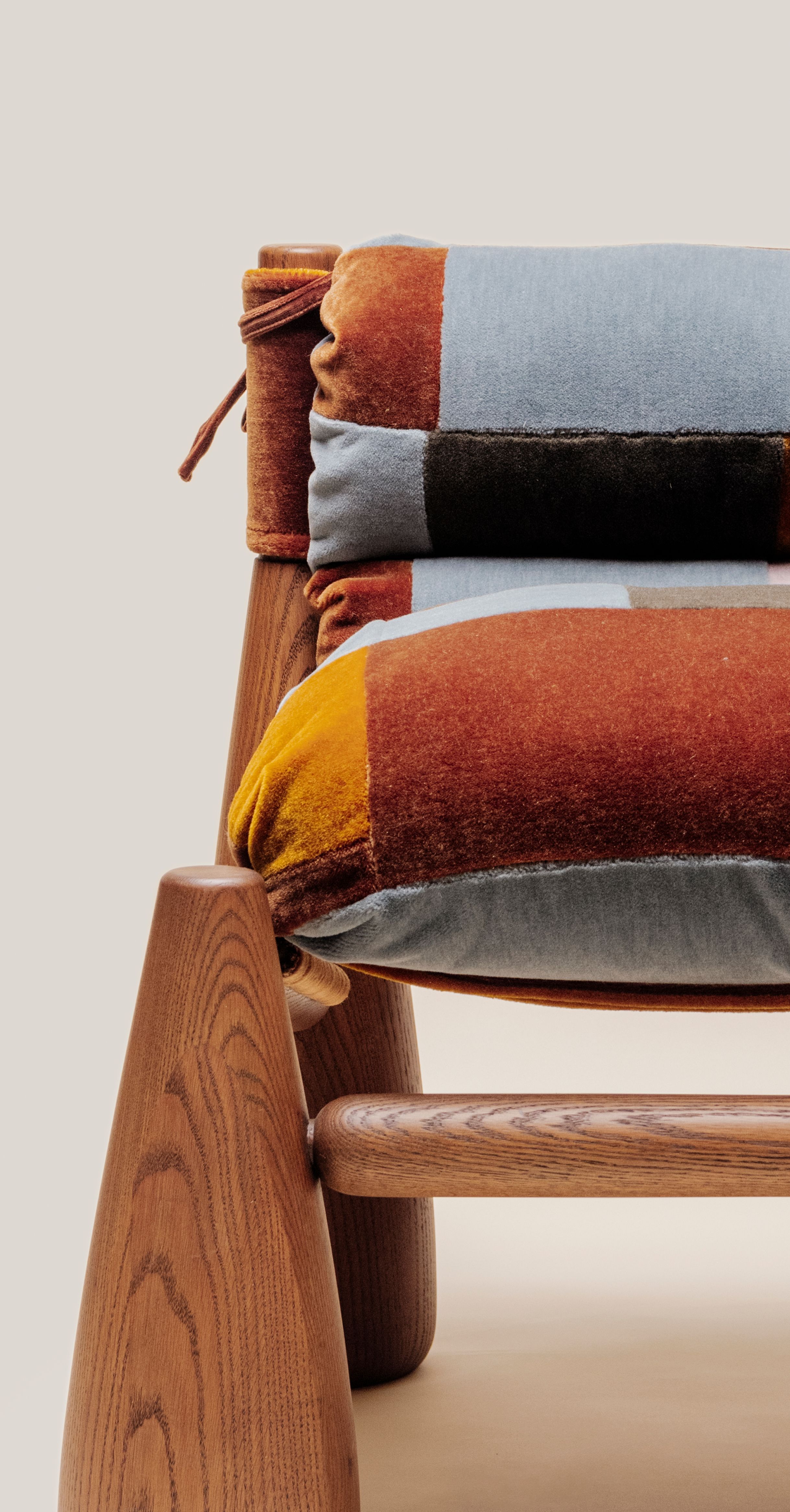 A detail image of a wooden armchair with sculpted cone-shaped legs and two thick, patchwork velvet cushions.