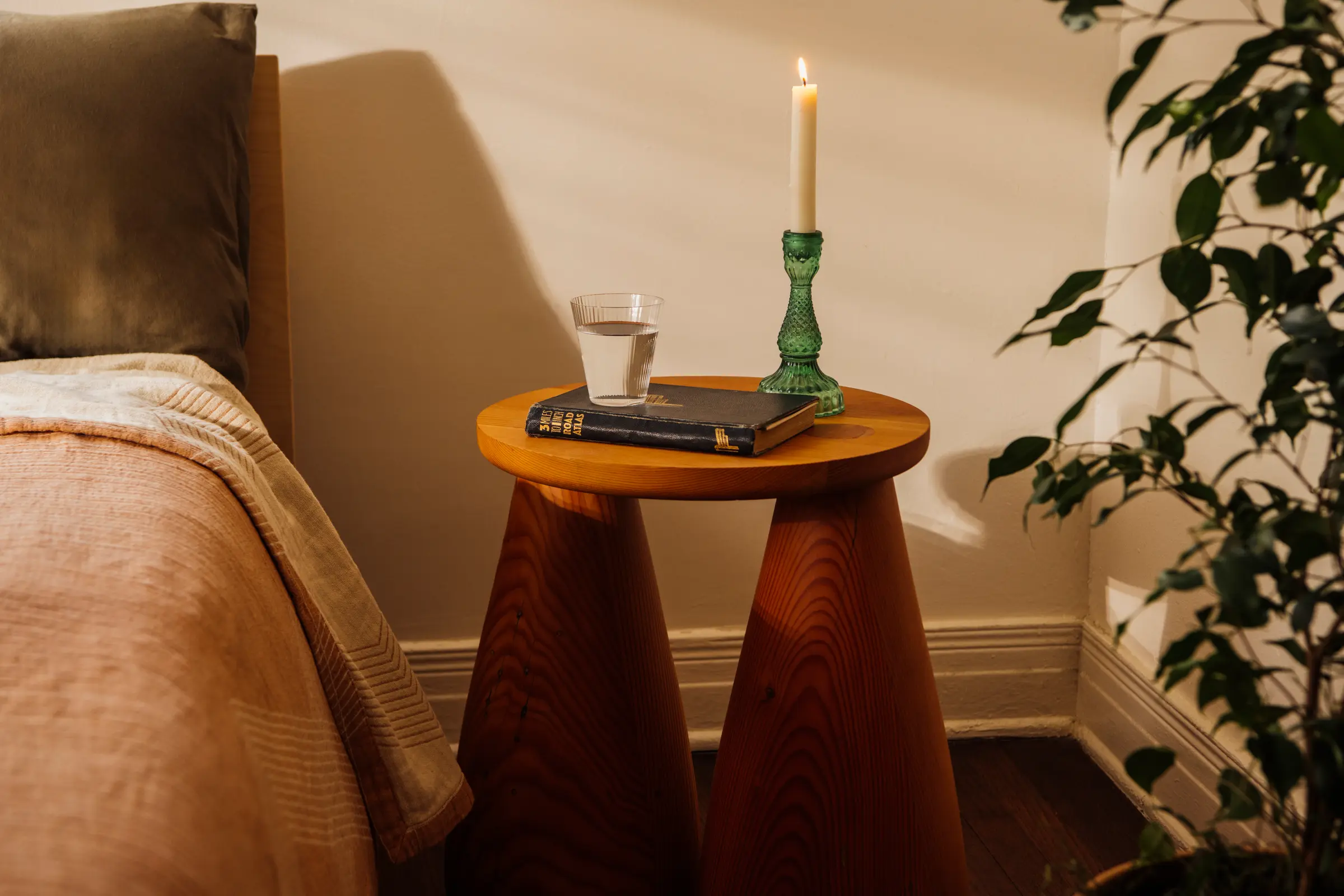 A wood table with bell legs and a book and candle and glass of water are placed on top, next to a bed and some plants.