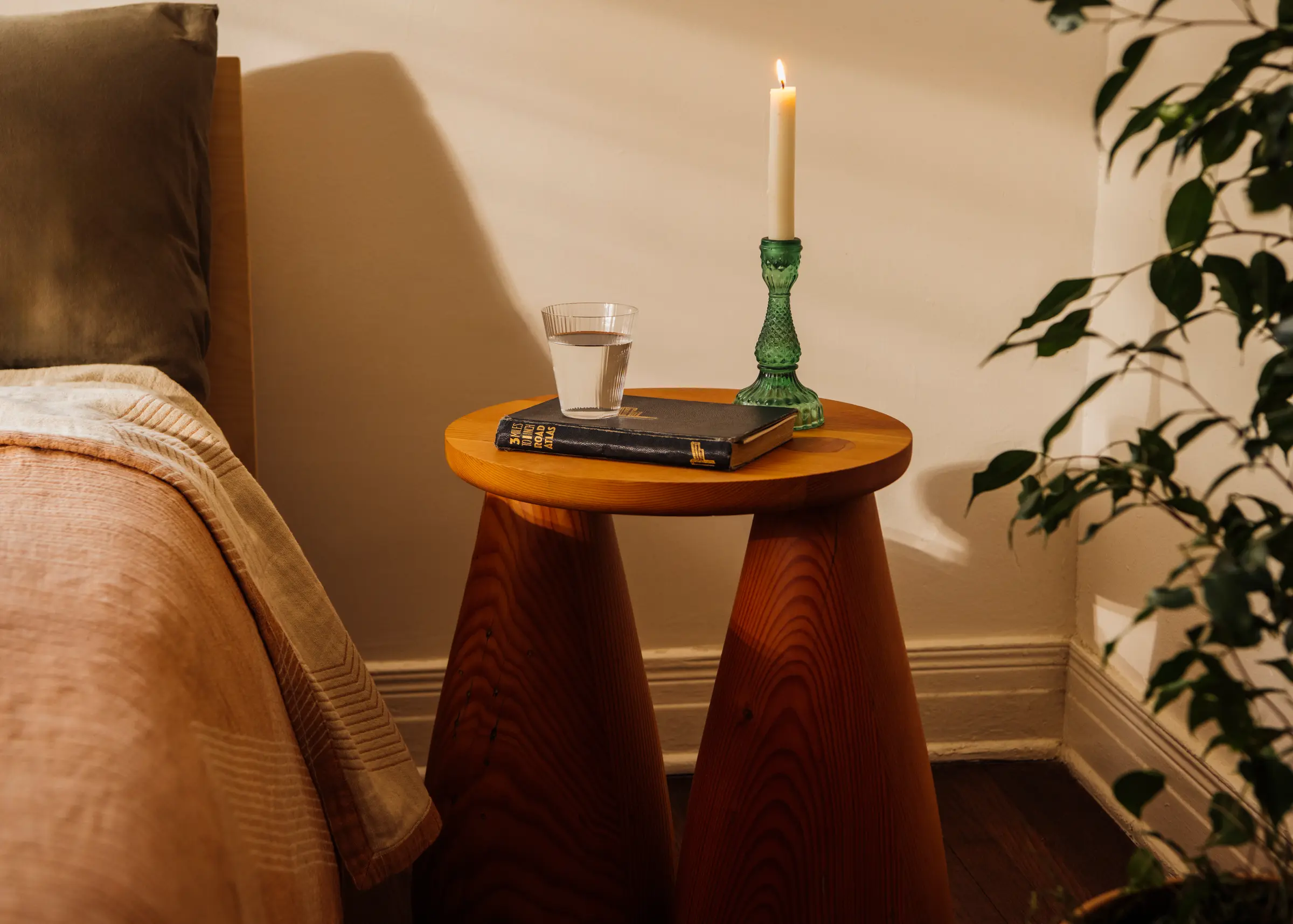 A wood table with bell legs and a book and candle and glass of water are placed on top, next to a bed and some plants.