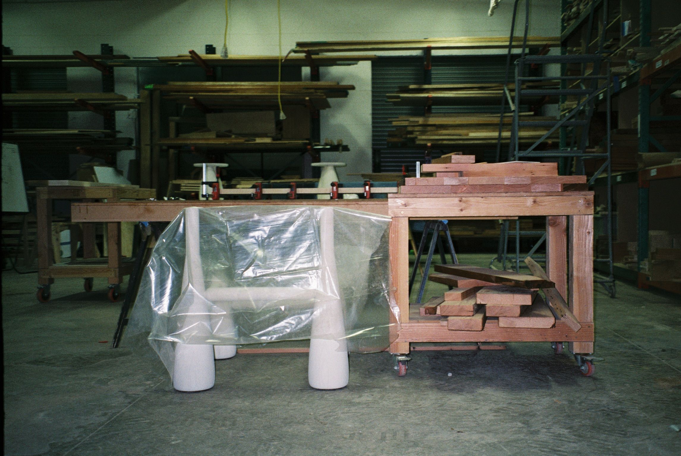 A wooden furniture frame with cone-shaped legs, partially covered in plastic, sits in a cluttered woodworking shop.