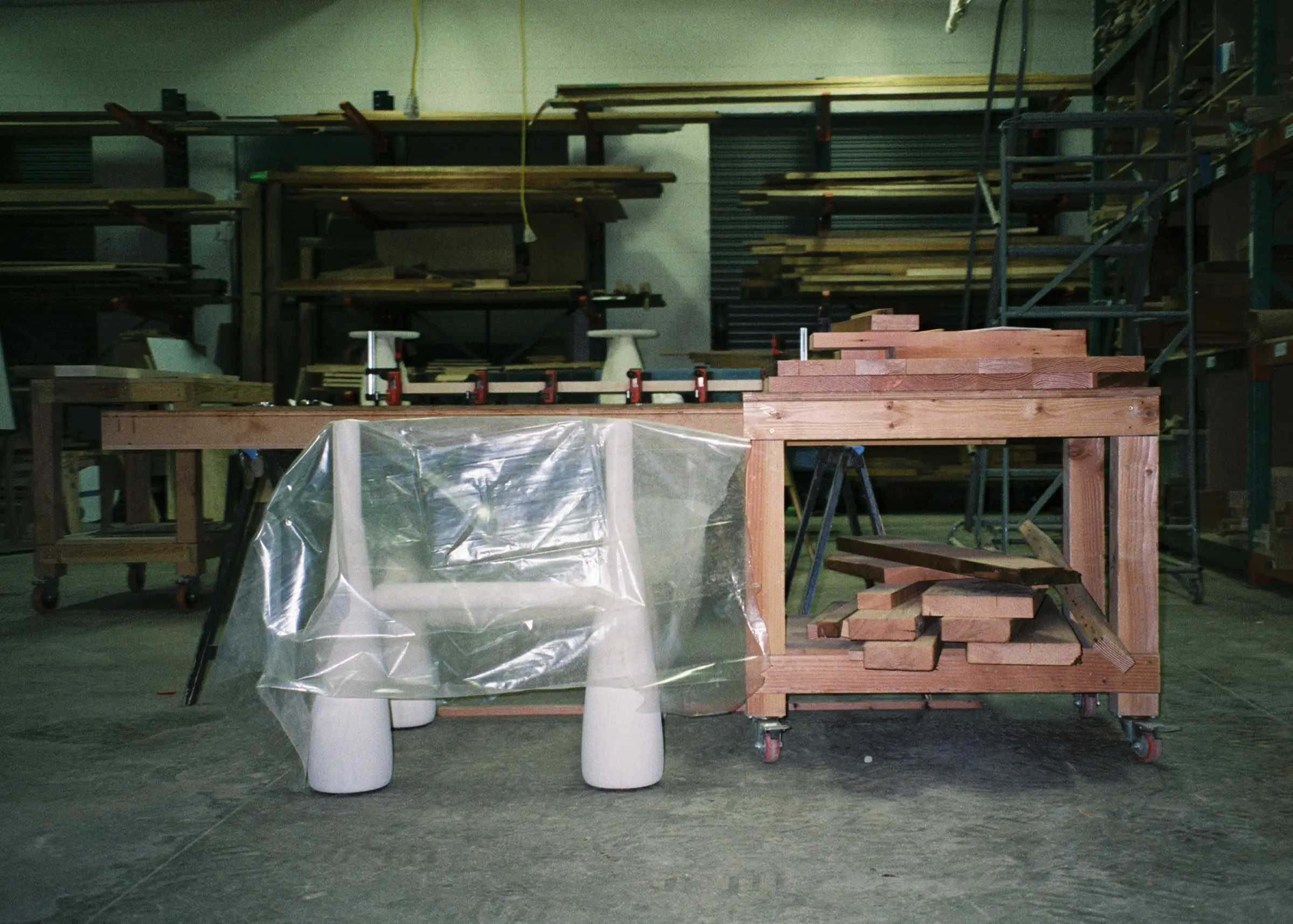 A wooden furniture frame with cone-shaped legs, partially covered in plastic, sits in a cluttered woodworking shop.