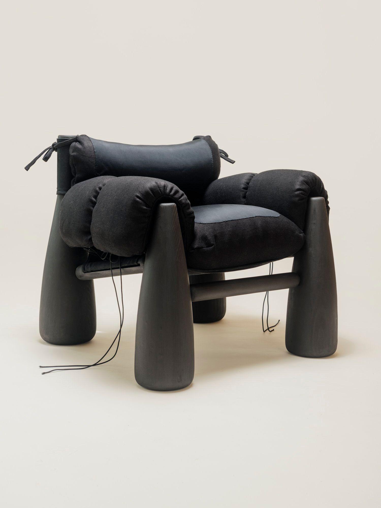 A three-quarter view of the Big Bell Chair, showcasing its modern and cozy design with a combination of black cushions and rounded black legs. The armrests and headrest are tied with black strings, emphasizing its contemporary, sculptural aesthetic.