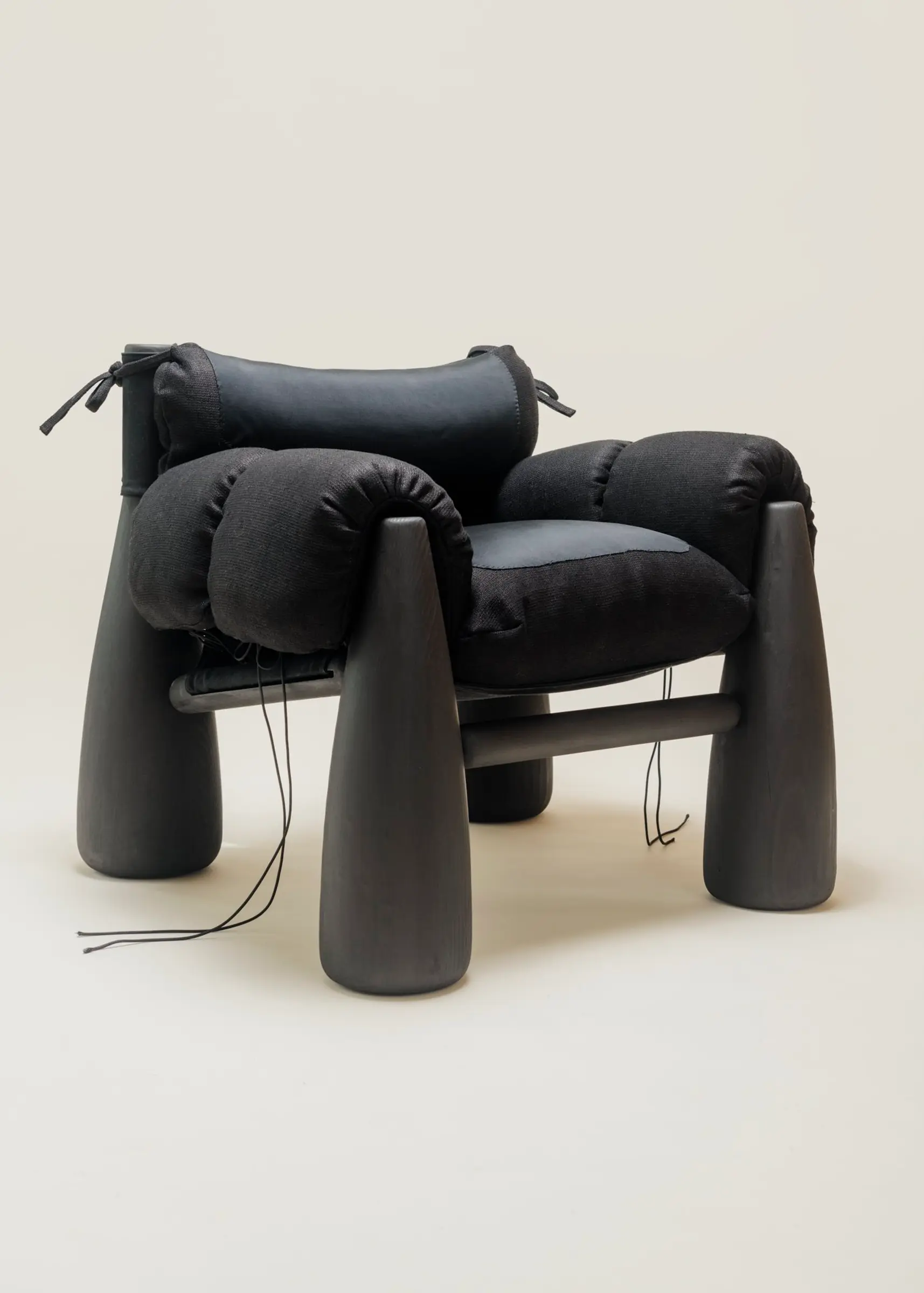 A three-quarter view of the Big Bell Chair, showcasing its modern and cozy design with a combination of black cushions and rounded black legs. The armrests and headrest are tied with black strings, emphasizing its contemporary, sculptural aesthetic.