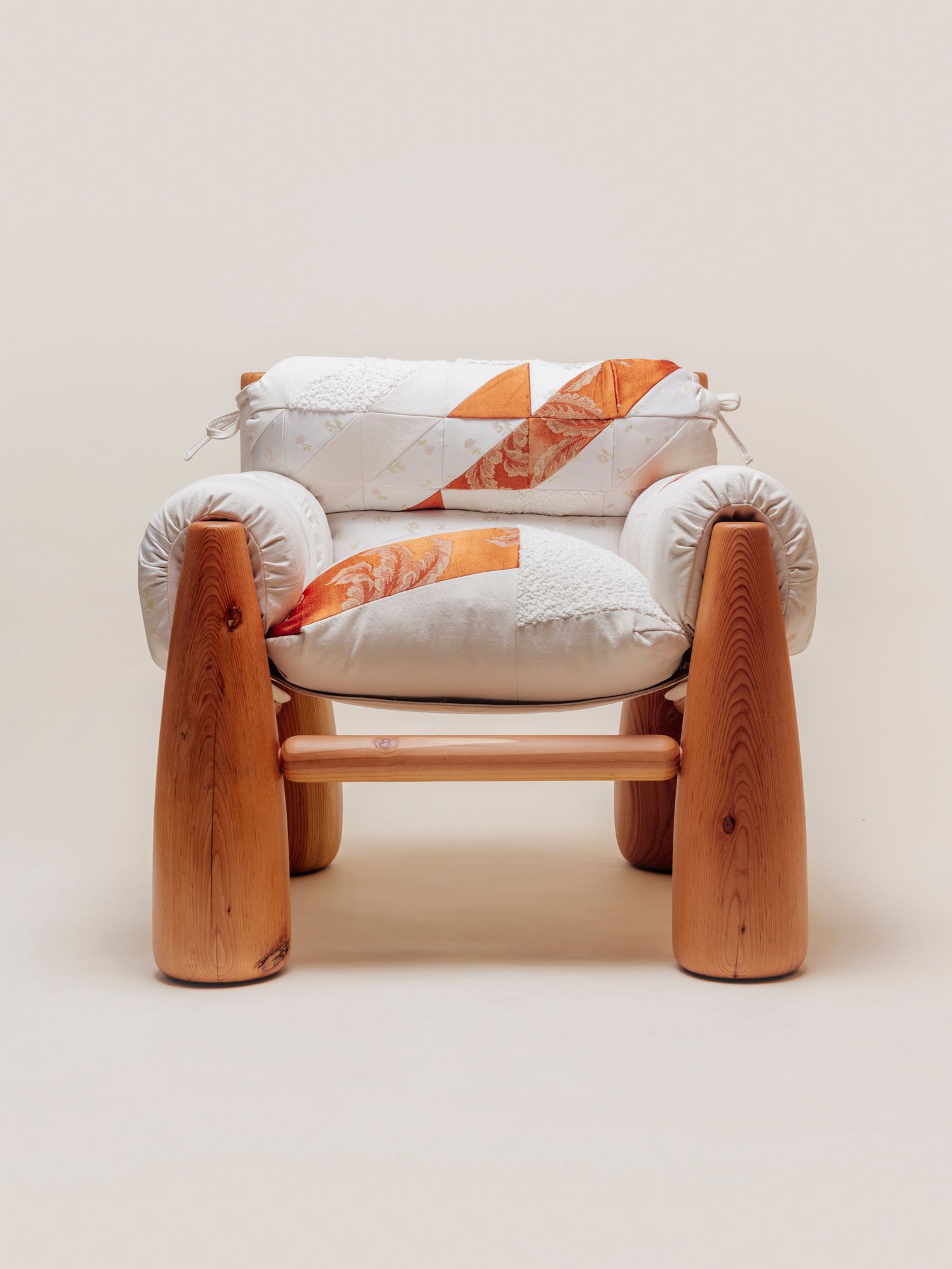 A cozy wooden chair with thick, rounded legs and quilted patchwork cushions in white and orange tones, viewed from an angle.