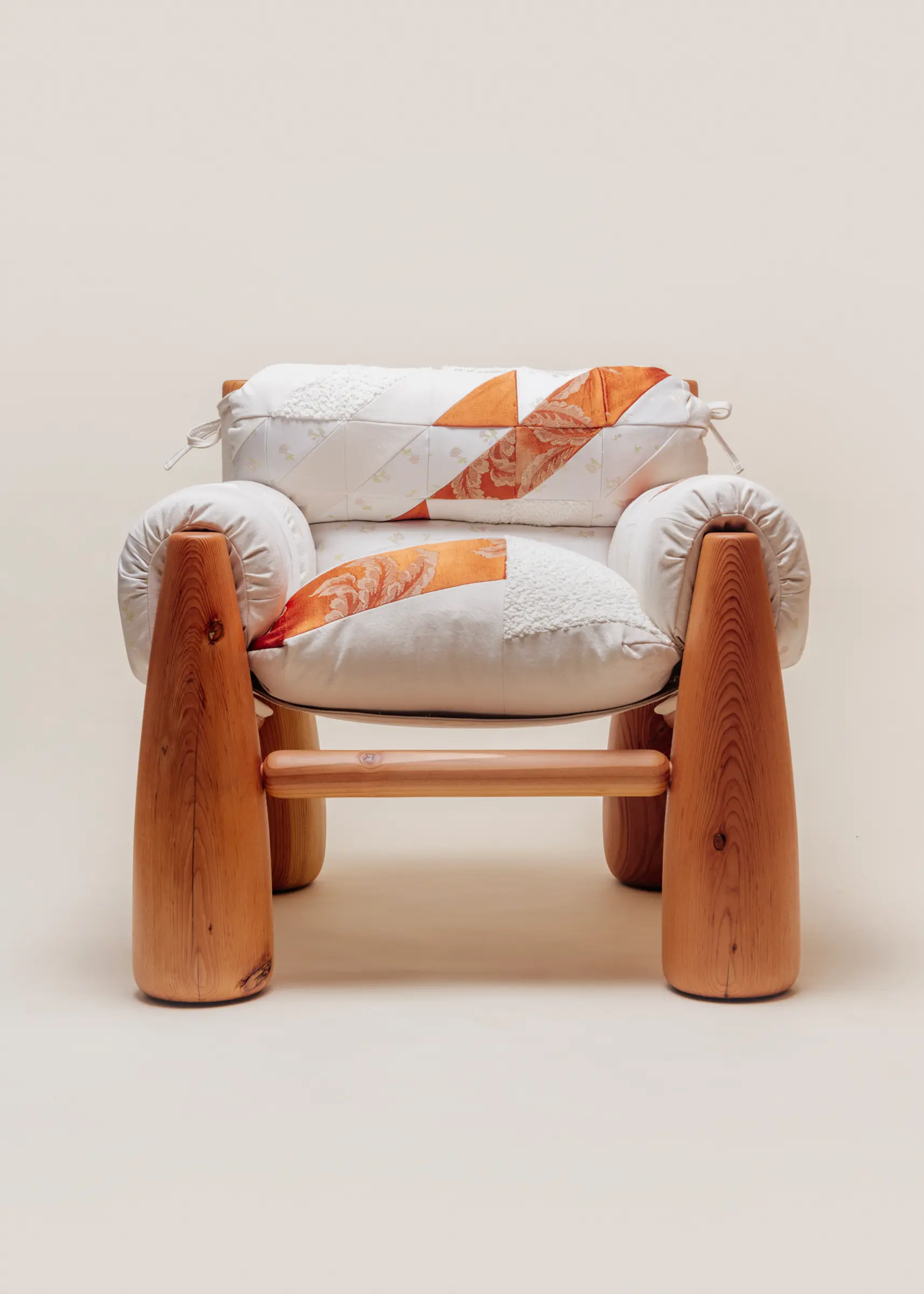 A cozy wooden chair with thick, rounded legs and quilted patchwork cushions in white and orange tones, viewed from an angle.