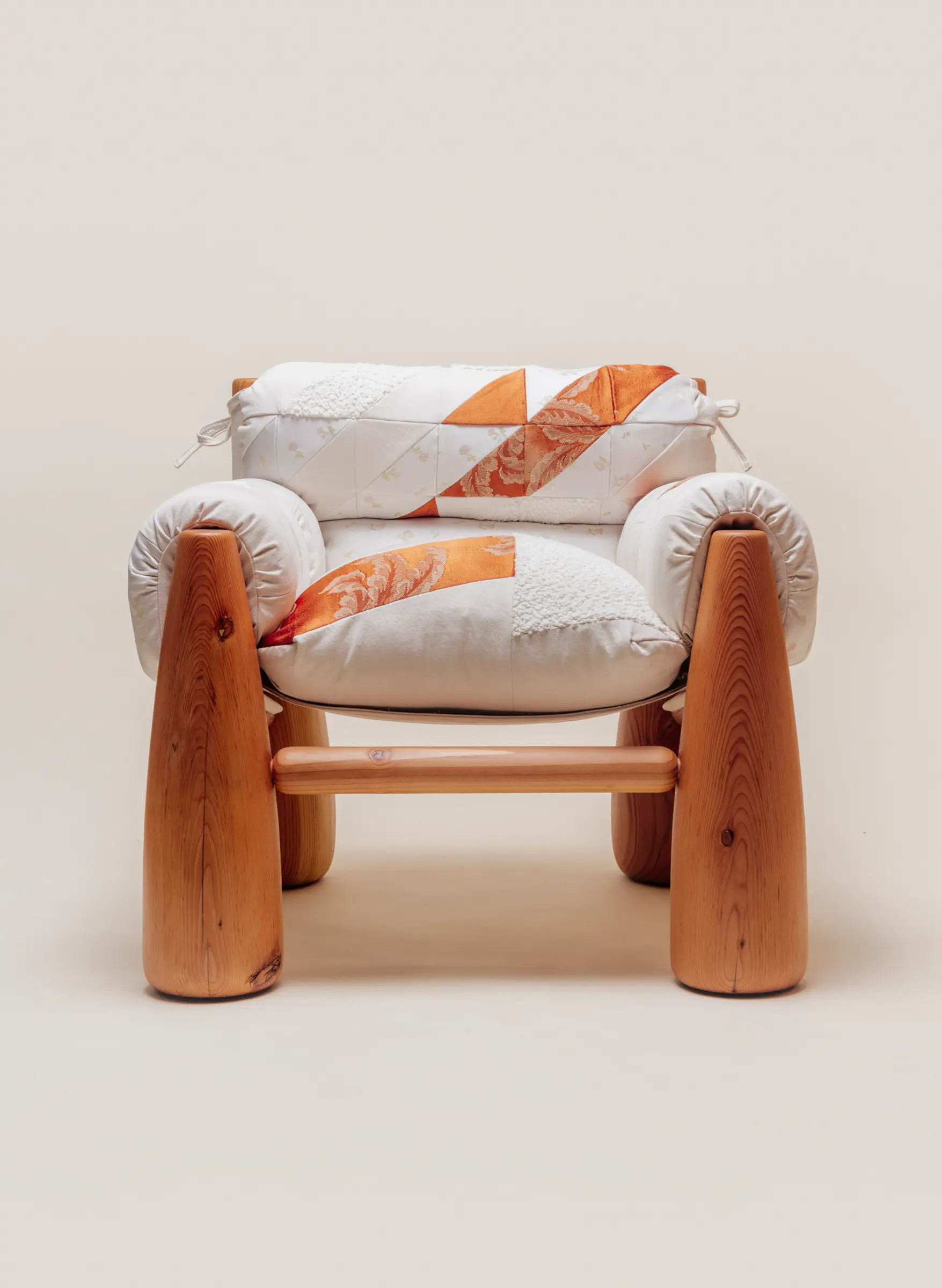 A cozy wooden chair with thick, rounded legs and quilted patchwork cushions in white and orange tones, viewed from an angle.