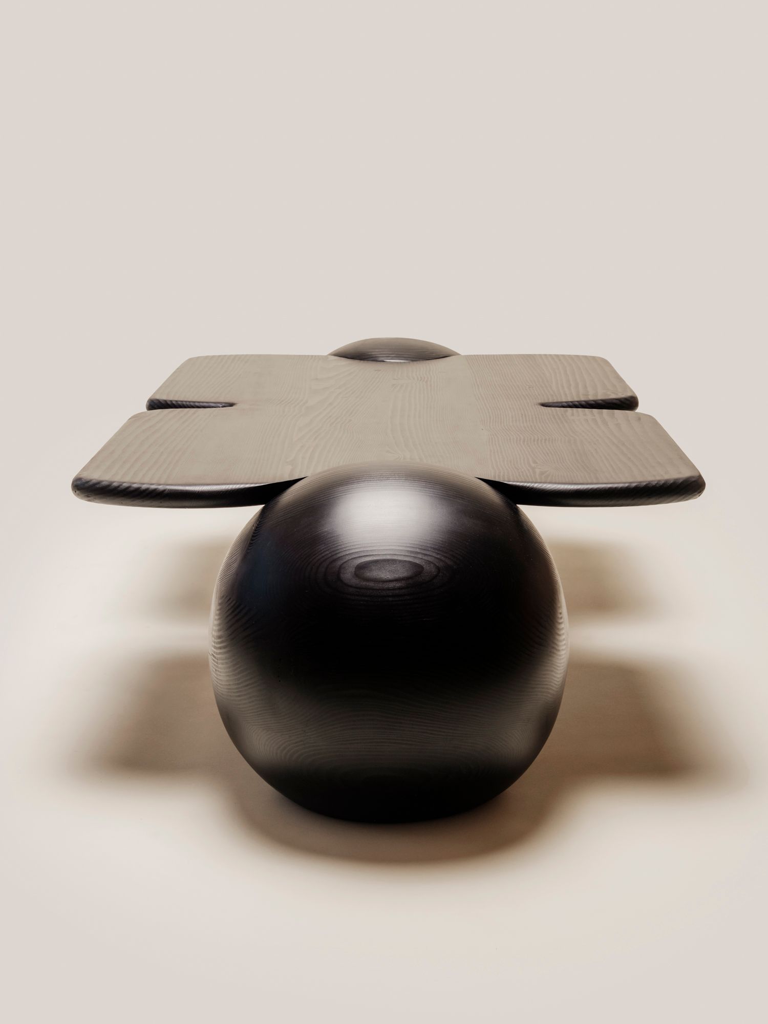 An end-on view of a sculptural black table, showing its cross-shaped top balancing on a large sphere.