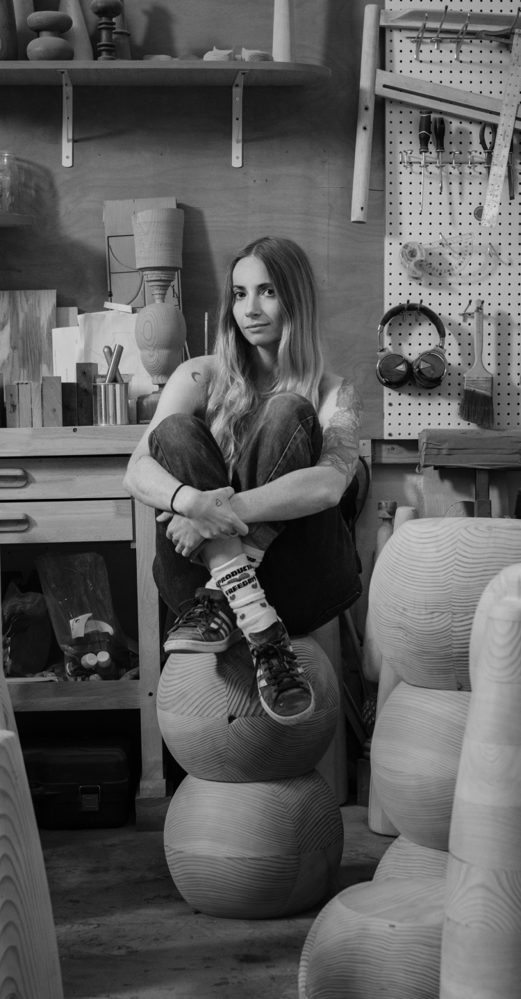 A black and white photo of Sam sitting amongst her sculptural wooden furniture in a busy workshop.