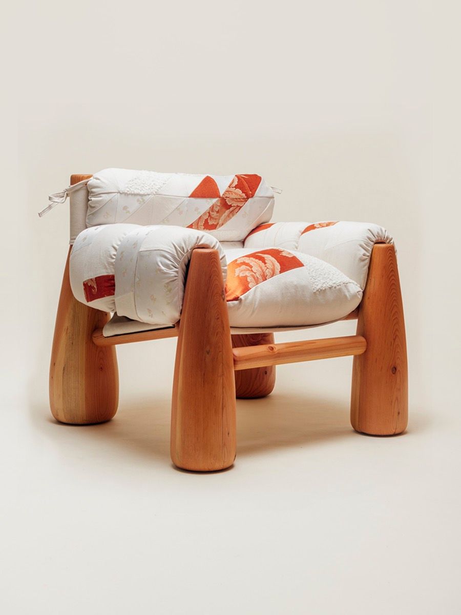A cozy wooden chair with thick, rounded legs and quilted patchwork cushions in white and orange tones, viewed from an angle.