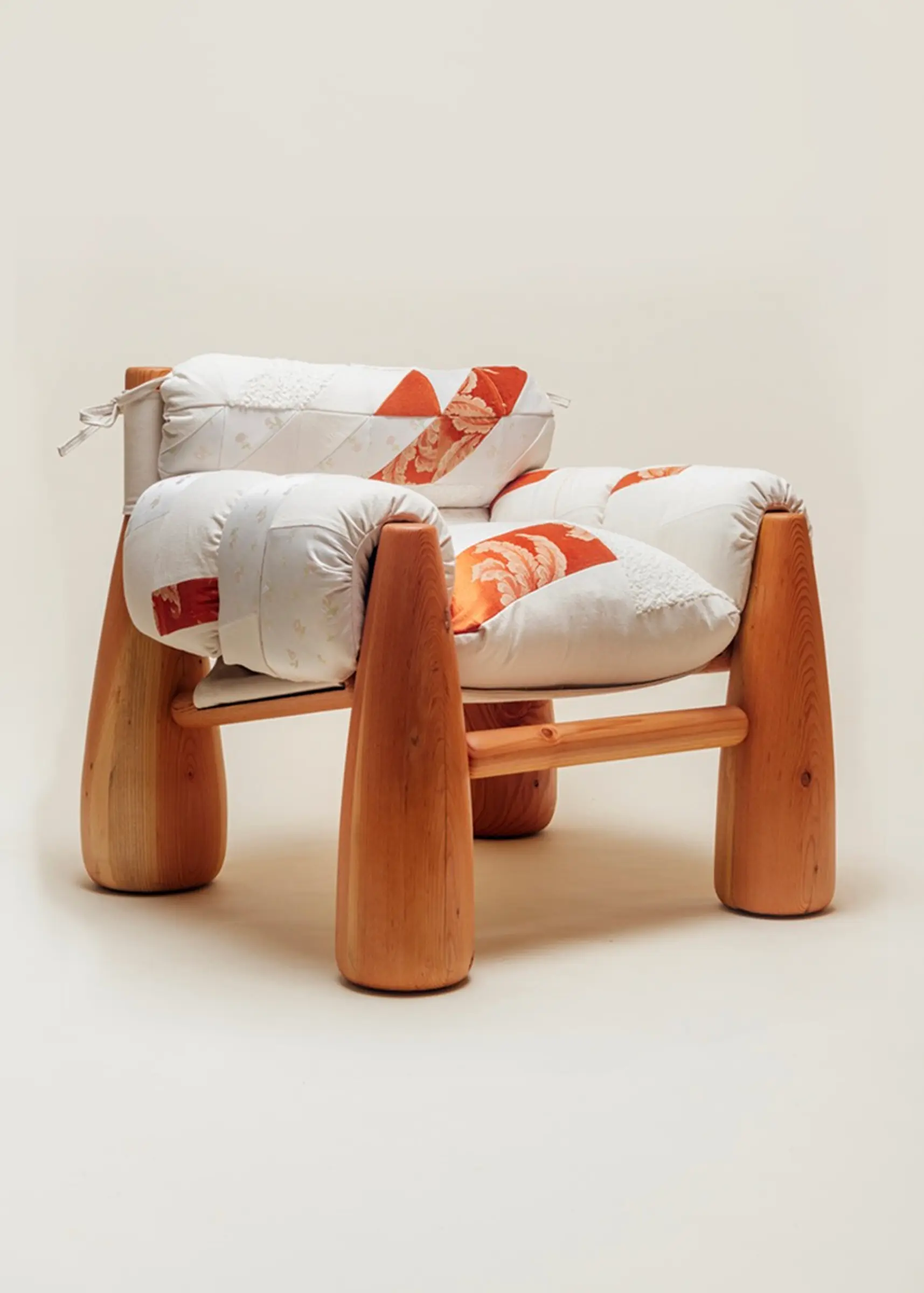A cozy wooden chair with thick, rounded legs and quilted patchwork cushions in white and orange tones, viewed from an angle.