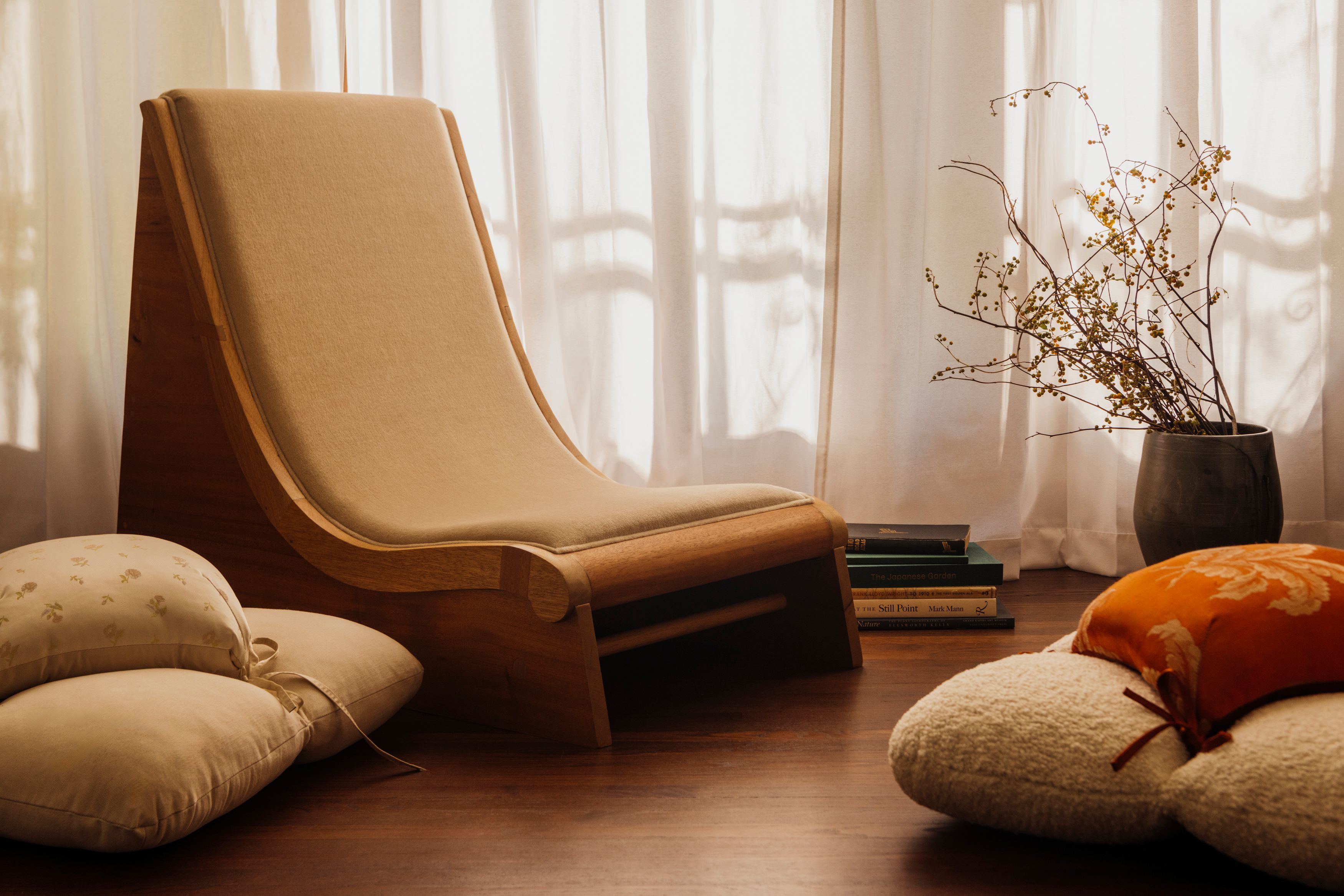 Chair placed next to cushions on the floor in front of a curtain with light shining through and a vase with flowers.