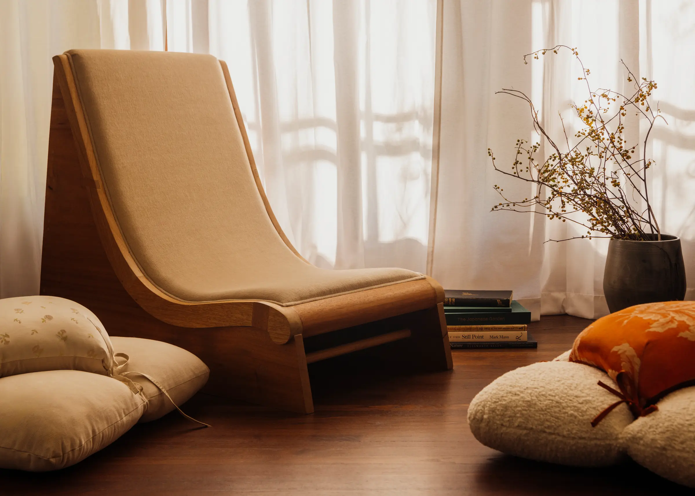 Chair placed next to cushions on the floor in front of a curtain with light shining through and a vase with flowers.