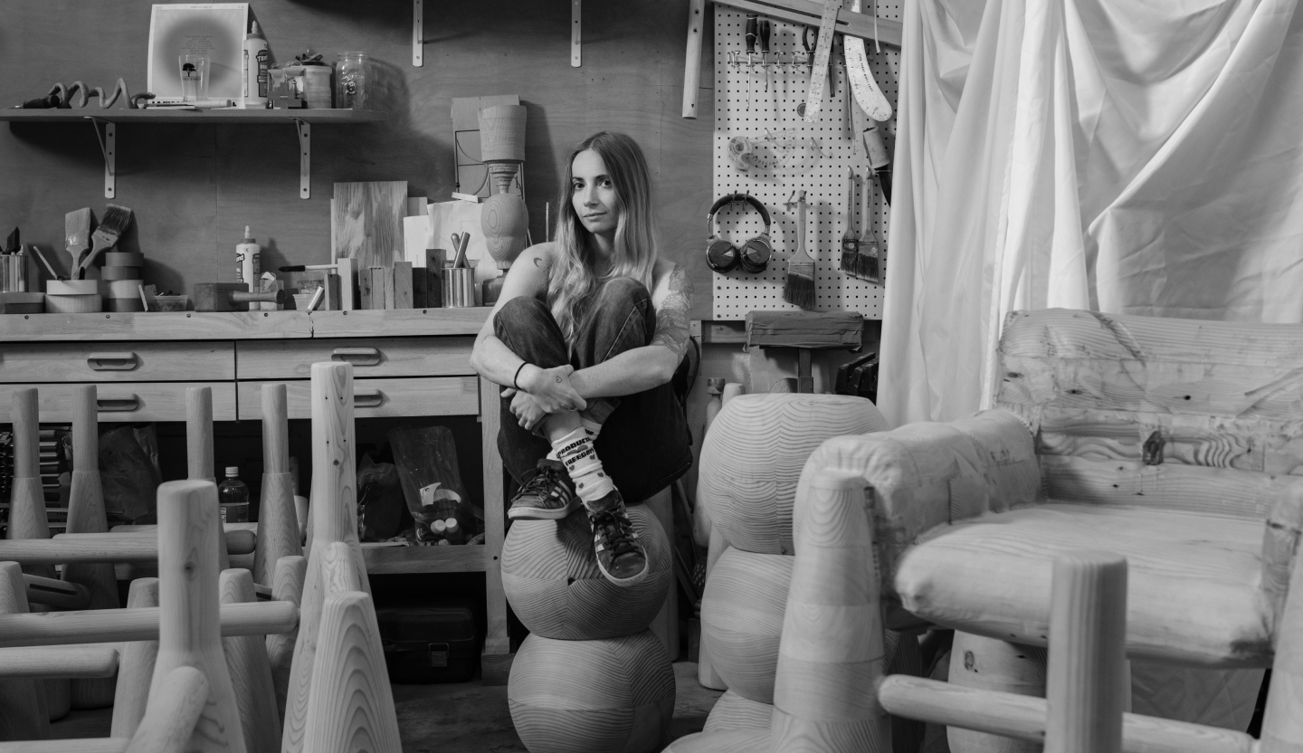 A black and white photo of Sam sitting amongst her sculptural wooden furniture in a busy workshop.
