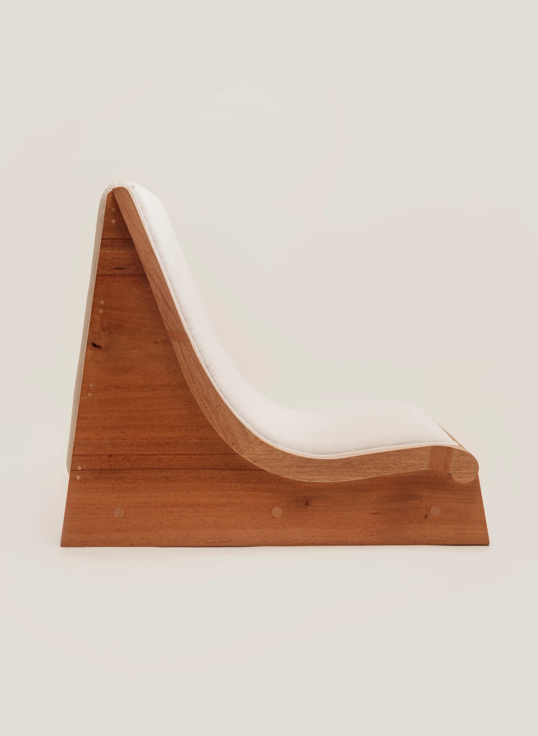 A side view of a low-slung wooden lounge chair with a single, continuous white cushion that follows the S-curve of the frame.
