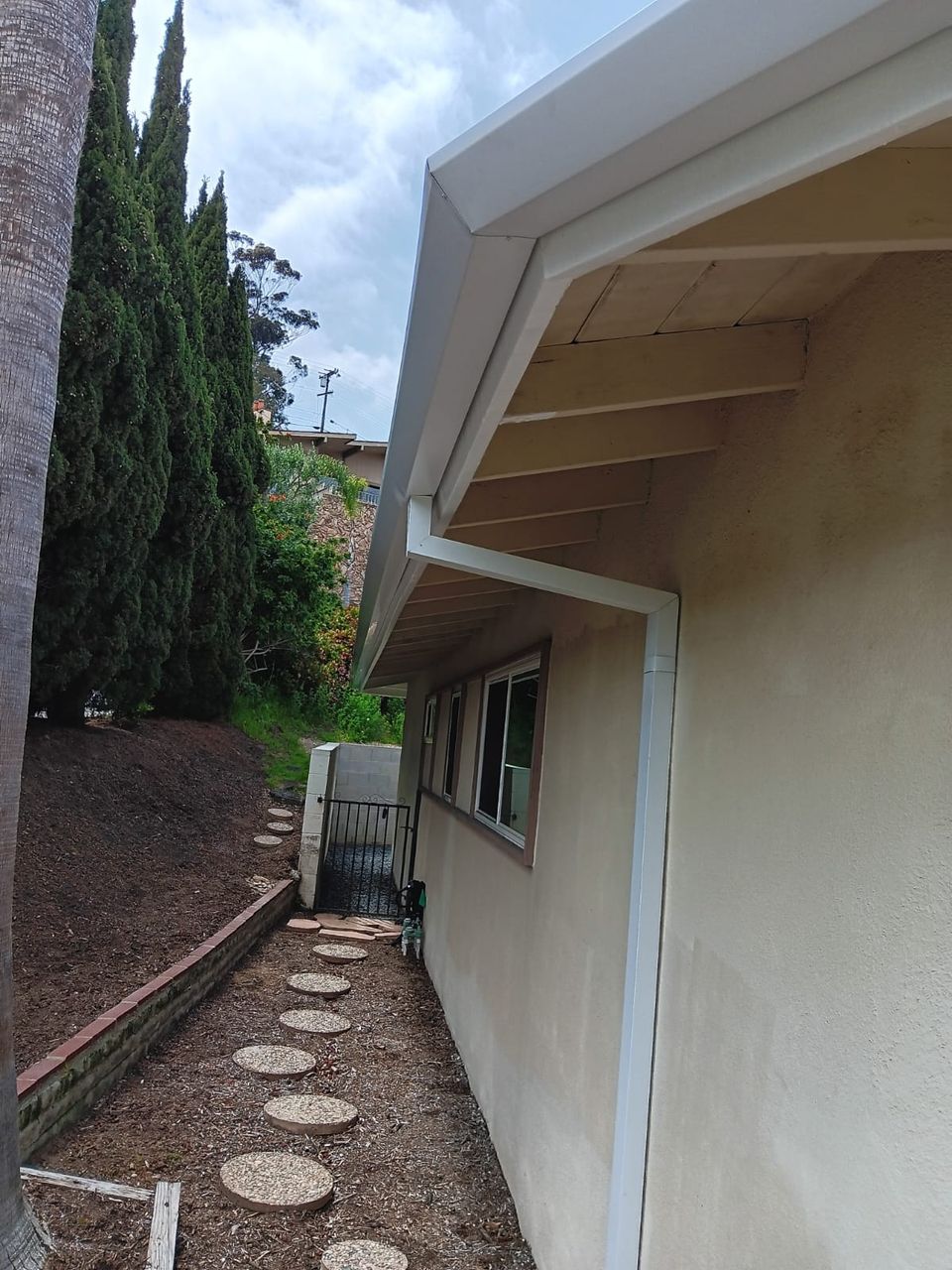White seamless gutter and downspout along a beige stucco home framed by cypress trees