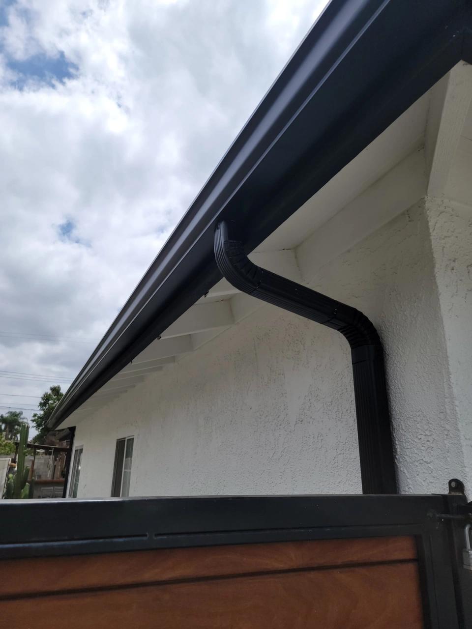 Black seamless gutter and downspout on white stucco beneath a dramatic cloudy sky