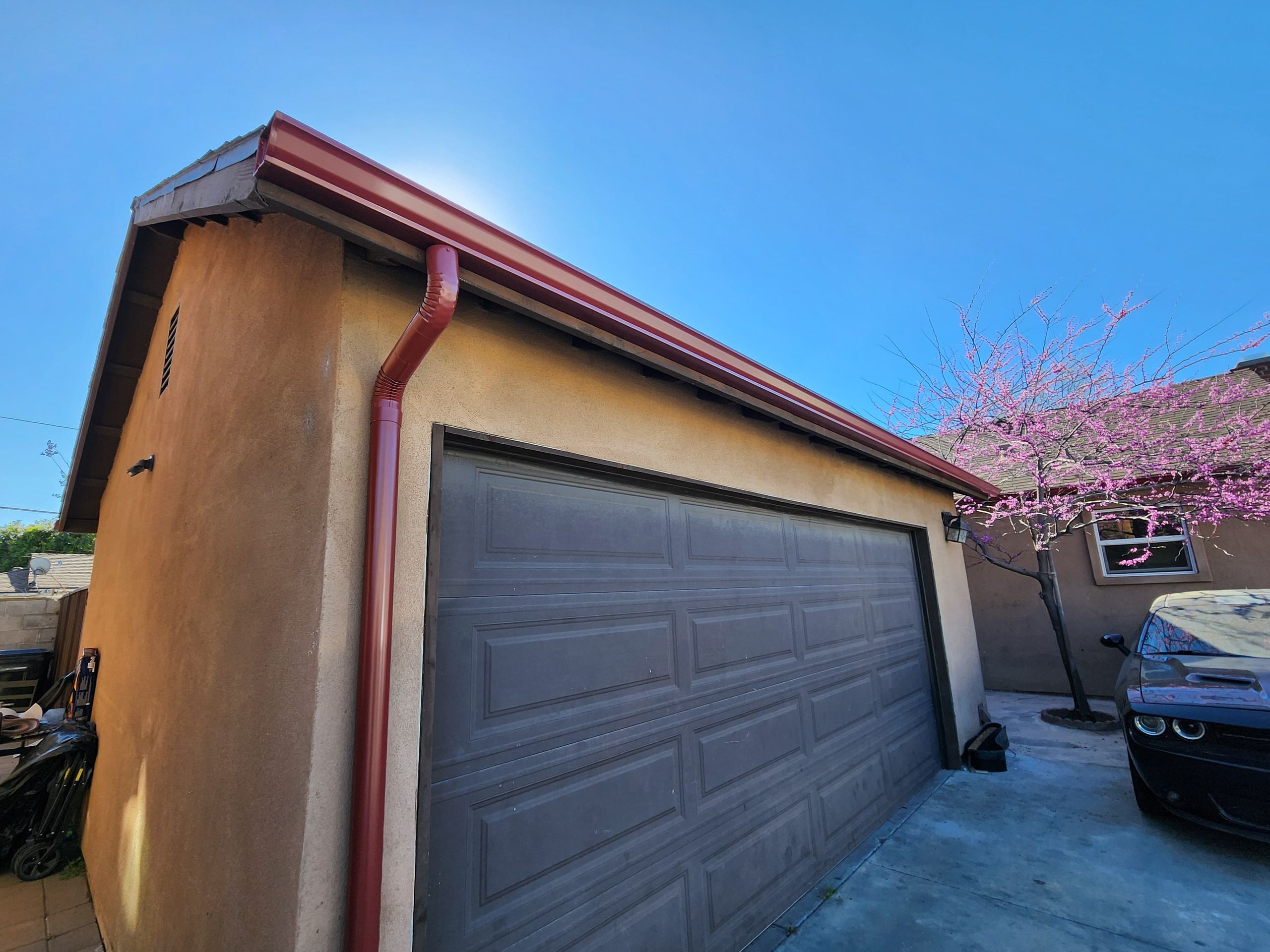 Burgundy seamless gutter and downspout on a tan stucco garage beside a redbud tree in bloom