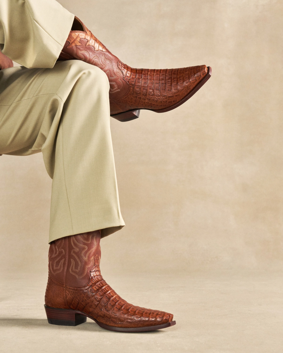 The Garrett in Pecan Hornback Caiman. A person wearing light beige pants and striking pecan brown textured Hornback Caiman cowboy boots sits with one leg crossed over the other against a plain background.