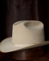 A light beige cowboy hat with a wide brim sits atop a wooden stand against a dark, textured background.