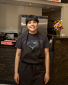 A person wearing a black "CLARIS" T-shirt and black apron stands smiling in a commercial kitchen setting with a flower vase on the counter behind them.