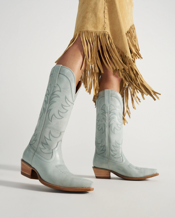The Annie in Slate Blue Cowhide. Person wearing light blue cowboy boots and a tan suede skirt with fringe.