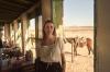 A woman in a cream peasant blouse stands on a ranch-house porch, with a saddled horse and desert hills behind her.