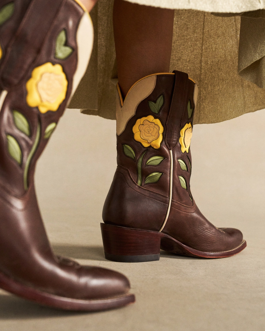 The Jolene in Truffle Cowhide. Dark brown truffle cowhide cowboy boots with yellow rose and green leaf appliqué, worn with an olive skirt. A taller matching boot is visible in the foreground.