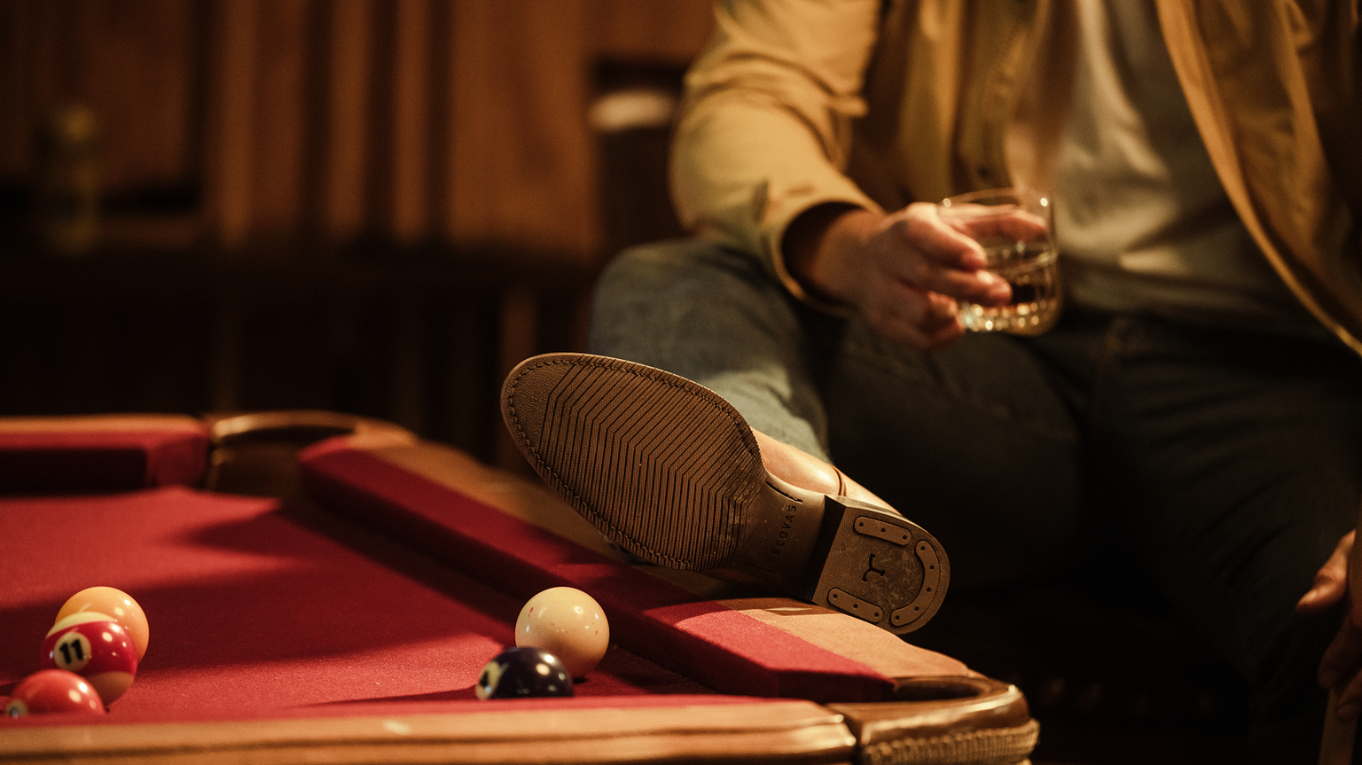 Person sitting on a pool table edge, holding a drink in one hand. Foot rests on table, and several pool balls are visible.