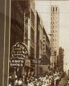 A busy city street with crowds, cars, and storefront signs including Bob's Loan Co., Lebo's Shoes, Kress, and Stein's. Tall buildings line both sides of the street.