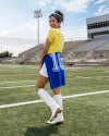 A young woman in a yellow top, white shorts, and white boots stands on a football field, smiling and looking over her shoulder. A blue shirt with the number "15" is tied around her waist.