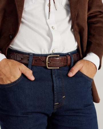 A person wearing a white shirt, brown jacket, and blue jeans with a brown leather belt, standing with hands in front pockets.