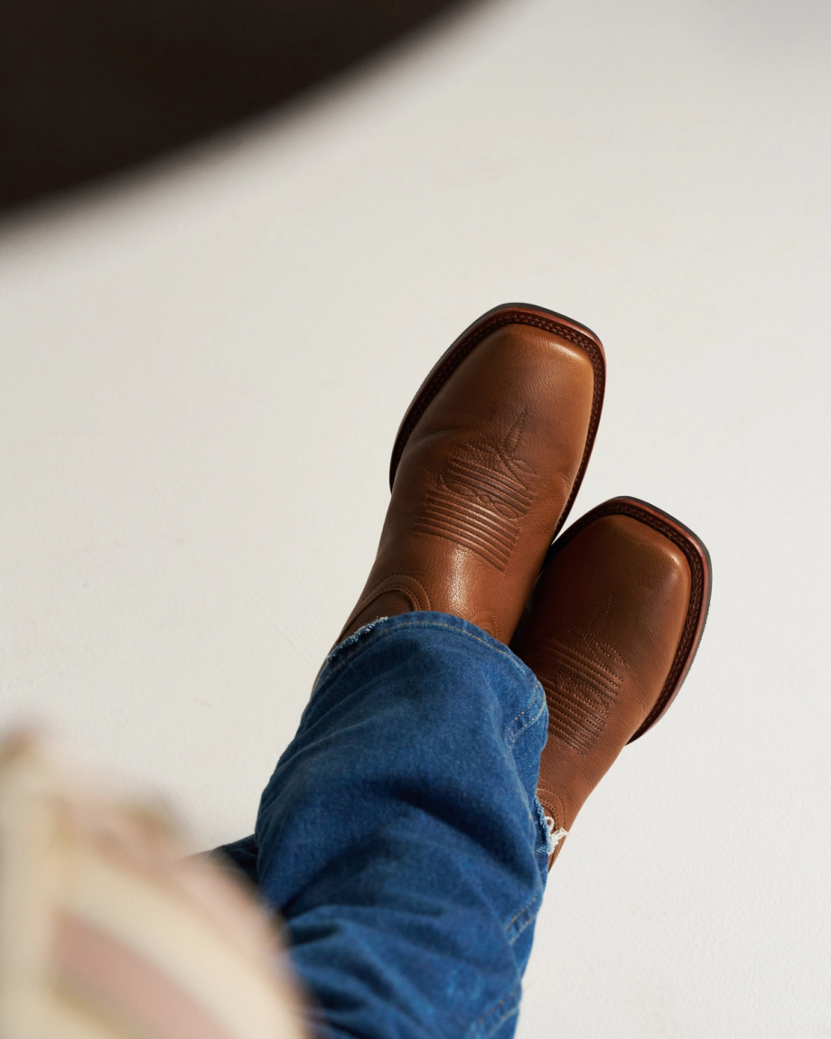 The Dusty in Mesa Goat. Person wearing blue jeans and brown leather cowboy boots, viewed from above against a plain light background, evokes the rugged charm of the Mesa, where even a wandering goat wouldn’t look out of place.