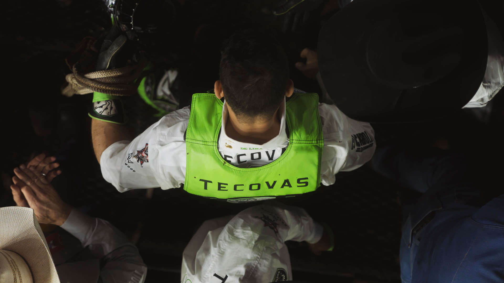 A person with "Tecovas" on their green vest is seen from behind, surrounded by others, some holding ropes.