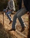 Two people wearing jeans and boots sit on a rusted metal fence in a grassy, rural outdoor setting. Their faces are not visible.