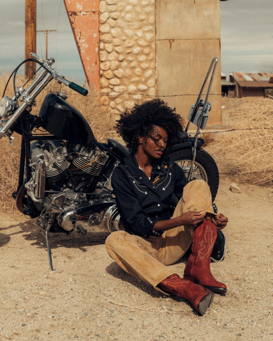 The Britt in Redwood Goat. A person wearing a black shirt, tan pants, and red boots sits on the ground next to a motorcycle in a rustic, desert-like setting, channeling the adventurous spirit of the Redwood Goat.