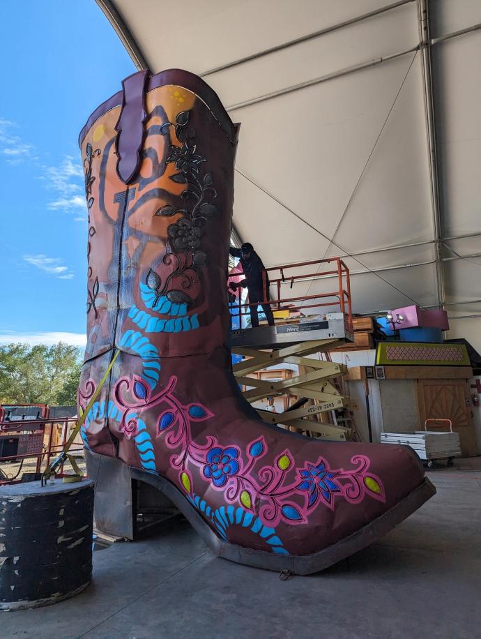 A large, colorful cowboy boot sculpture with floral designs being painted by an artist on a scaffold.