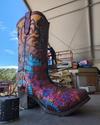 A large, colorful cowboy boot sculpture with floral designs being painted by an artist on a scaffold.