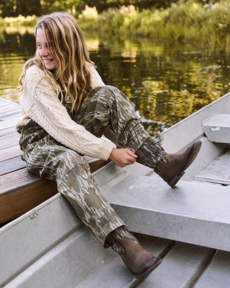 A person with long hair in a sweater and camouflage pants sits on a dock by the water, smiling and adjusting their brown boots while sitting on the edge of a metal boat.