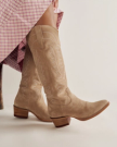 Person wearing tan suede cowboy boots with white stitching and a mid-heel, paired with a pink plaid skirt.