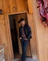 A man in a cowboy hat and casual western attire leans against a doorframe of a wooden building, looking to the side.