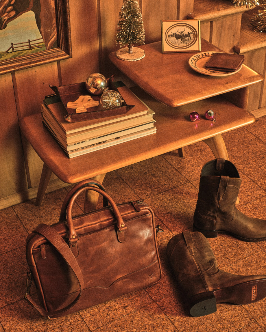 A wooden side table with books, ornaments, and decor sits in a room with a framed painting, leather bag, and brown boots on the tiled floor.