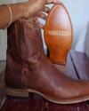A hand holds up a brown leather Tecovas cowboy boot to show the sole, with another boot resting on a wooden surface nearby.