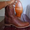 A hand holds up a brown leather Tecovas cowboy boot to show the sole, with another boot resting on a wooden surface nearby.