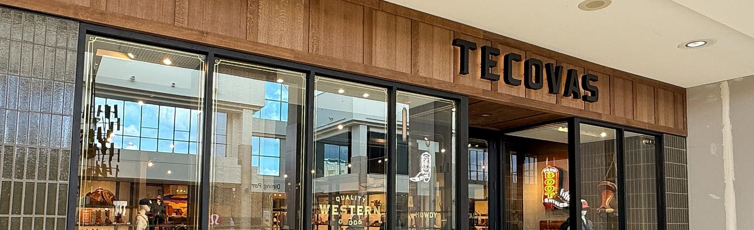 Exterior of a Tecovas store in a shopping mall, showing large glass windows and a wooden sign above the entrance.